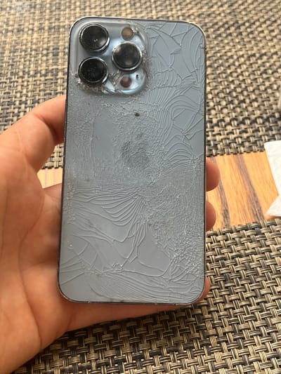 A hand holds a cracked iPhone 13 Pro Max phone. The phone's back is a light blue color, and it has a camera module with three lenses in the upper left corner. The screen of the phone is heavily cracked with a web-like pattern. The background appears to be a woven placemat on a wooden table. The image was taken in Otopeni, Romania.