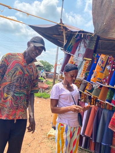 The image depicts an outdoor market scene in Amaseri, Nigeria, on a cloudy day. Two individuals are the central focus. On the left, a man wearing a black baseball cap and a brightly patterned, colorful shirt with swirling designs stands looking at the camera and smiling. His right arm is bent at the elbow, and his hand is partially visible. He is wearing black trousers. To the right of the man, a woman wearing a black headscarf and a light purple t-shirt is looking down at her hands, which are holding something small. She is wearing a colorful striped skirt with yellow, blue, red, and white patterns. A black crossbody bag is slung over her shoulder. Behind them, fabric stalls are laden with vibrantly colored and patterned textiles, displayed hanging vertically. The ground is a reddish-brown dirt path. In the background, other market elements are visible, including a wheelbarrow, some trees, and a glimpse of a vehicle. The overall environment suggests a bustling, open-air marketplace.