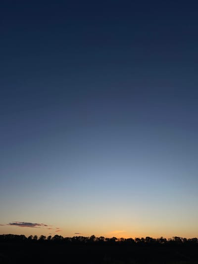 The image depicts a horizontal view of the sky at dusk or dawn. The left side of the image shows a deep blue sky, transitioning to a lighter blue and eventually to a light orange near the right edge. The horizon line is defined by a dark silhouette of trees. The image does not provide sufficient information to confirm or deny that it was taken in the city of Popasne, Ukraine. No people, actions, or interactions are visible in the image.