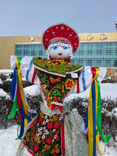 A large, traditional Russian effigy, approximately human-sized, stands upright amidst snow-covered ground and shrubs. The effigy features a fabric head with painted blue eyes, red lips, and white pom-pom hair. It wears a prominent, ornate red kokoshnik-style headdress adorned with silver trim, red gemstones, and silver embellishments. The effigy's body is dressed in multiple layers of patterned fabric, including a colorful shawl with a black background featuring birds, flowers, and foliage, and a skirt with a small floral pattern, overlaid with an apron-like piece depicting red strawberries and white flowers on a black background. Its extended arms are constructed from straw or sticks, covered in white fabric, with multiple colorful satin ribbons (blue, red, yellow, green, and white) tied to both sides.
In the immediate foreground, dark green shrubs are visible, partially covered by white snow. The background features a large, modern building with a light beige or yellow facade and numerous blue-tinted reflective windows. Two large, white ring-shaped fixtures are visible on the building's roofline. The sky above is a clear, light blue. The scene is located in the city of Plast, Russia, under winter conditions.