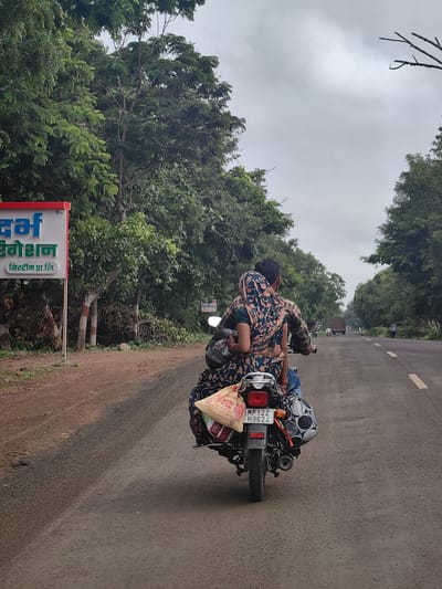 The image depicts a cloudy day in Motala, India. Two people are riding on a motorcycle, with the driver in front and a passenger behind. The passenger is wearing a colorful patterned sari. They are traveling on a paved road lined with trees. On the left side of the road, a red and white sign is visible with text in an Indian script. The motorcycle has bags attached to the back, suggesting they are carrying belongings. In the distance, other vehicles and a person are visible on the road. The overall environment appears to be rural or suburban, with lush greenery.
