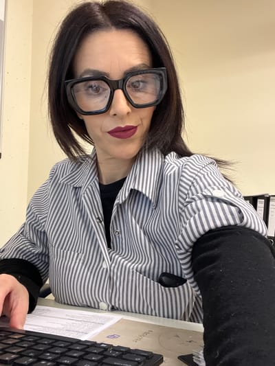 A woman with dark hair, wearing large black-framed glasses, a striped shirt, and dark lipstick, is photographed from a close, high-angle perspective. She is positioned at a desk, partially obscured by a black keyboard. A document is visible on the desk. The background is a plain beige wall, with binders visible in the upper right corner. Location details are not discernible in this image, therefore I cannot attest that this image was taken in Mensdorf, Luxembourg.