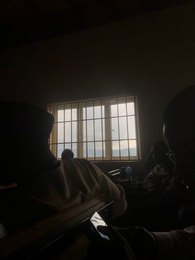 The image depicts an indoor scene in Farin Yashe, Nigeria, dimly lit with light streaming through a barred window. In the foreground, a person is partially visible, silhouetted as they hold a smartphone. To their left, another person is silhouetted, facing away from the camera and towards the window, wearing a light-colored garment. Beyond them, further into the room, more individuals are dimly visible in the shadows. The window reveals a muted, overcast sky and the faint outlines of hills or mountains in the distance. On a surface near the window, a bottle of water is present. The overall atmosphere is one of quiet observation or study, with the limited light and the presence of individuals engrossed in their own activities.