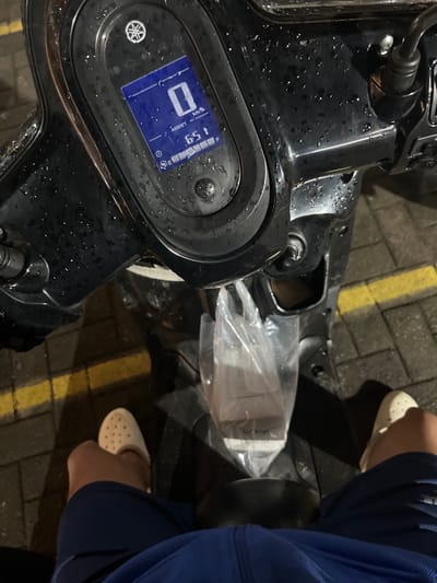 The image shows a close-up view of a person sitting on a black scooter. The scooter's digital dashboard is visible, displaying "0 km/h" and the time "6:51". The dashboard and surrounding areas of the scooter are speckled with water droplets, indicating it might be raining or has recently rained. A clear plastic bag containing what appears to be a beverage is hanging from the handlebars, resting on the seat. The person is wearing blue shorts and light-colored slip-on shoes. The background shows a brick-paved ground with a yellow line, typical of a street or parking area. The scene suggests the person is stopped on their scooter, possibly waiting or having just arrived. The location is identified as Boja, Indonesia.