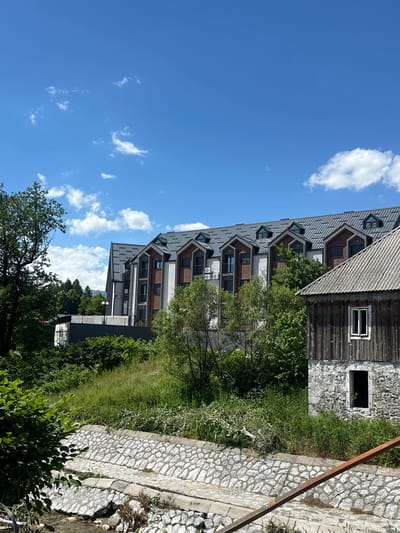 The image depicts a view of Dulovine, Montenegro, featuring a modern apartment building with a dark grey tiled roof, white and brown facade, and multiple balconies, situated behind a stone-lined canal. To the right of the apartment building, an older, rustic stone and wood structure with a corrugated metal roof and a small window is visible. Lush green vegetation, including trees and grass, surrounds the buildings and the canal. The sky is a bright blue with scattered white clouds. The scene appears tranquil and is captured on a sunny day. There are no people or animals present in the image.