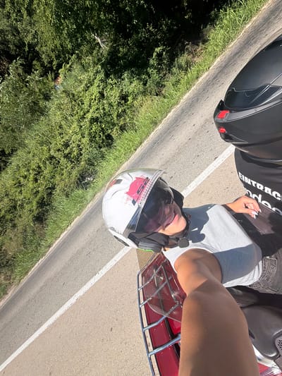 In Mišići, Montenegro, on a sunny day, two individuals are riding on a motorcycle. The person in the foreground, wearing a white helmet with red accents and a grey tank top, is smiling and looking towards the camera. They have fair skin and white painted fingernails. The second person, positioned behind the first, is wearing a black helmet and a black t-shirt with white text that appears to read "EINTRACHT". The motorcycle has a red luggage rack. The scene is set on a paved road with a white line marking the lane, bordered by lush green vegetation and trees on the left side of the road. The perspective is from a high angle, looking down at the riders.