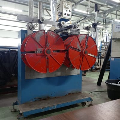 The image depicts an industrial setting, likely a workshop or factory, with a prominent piece of machinery in the foreground. The machine features two large, red, spoke-like spools mounted on a blue base. Above the spools, there is a metal frame with several rollers and a control box with buttons and indicators. The machine appears to be used for winding or unwinding material. In the background, there are industrial pipes, lighting fixtures, and other equipment. A person can be seen partially in the frame on the left, wearing dark pants and sandals, standing near the machine. To the right, a black bucket filled with white material, possibly cable or wire, is visible. The floor is concrete, and the overall environment suggests an active industrial workspace. There are no specific visual cues in the image that would indicate the location as Kinel, Russia.