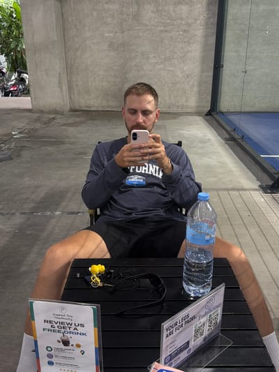 A fair-skinned man with short brown hair and a beard sits on a black chair with his legs spread apart, looking at his phone. He is wearing a dark blue long-sleeved shirt with a white and blue graphic on the chest that reads "CALIFORNIA USA" and black shorts. On the black table in front of him, there is a bottle of water, a yellow keychain with a duck figure, black earphones, and a sign that reads "Support Our Community REVIEW US & GET A FREE DRINK." Another sign on the table says "YOUR LEGS ARE FOR PADEL. Padelicious". The man is at an outdoor facility with a concrete floor and a concrete wall behind him. To the right of the man, there is a blue padel court with glass walls. In the background to the left, motorcycles are parked near some lush green foliage. The lighting suggests it is daytime.