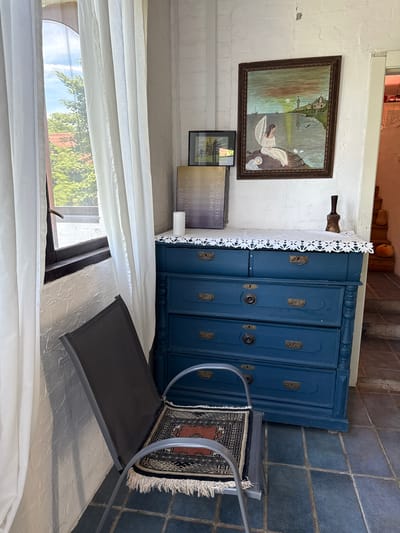 In Alagir, Russia, a corner of a room is depicted with a window on the left, covered by white curtains. Through the window, trees and a red-tiled roof are visible under a blue sky. A textured white wall is present. In the center of the image is a blue dresser with ornate brass drawer pulls. A white doily or tablecloth with an intricate pattern adorns the top of the dresser. On the dresser sits a white candle and two framed pictures, one larger with a painting of a woman on a rock by the sea and a smaller one with a depiction of a building. To the right of the dresser, a staircase with terracotta-colored risers and concrete steps leads upwards. Two pumpkins are visible on the staircase. In the foreground, a dark grey metal chair with a patterned rug on its seat is positioned in front of the dresser. The floor is covered with blue square tiles.