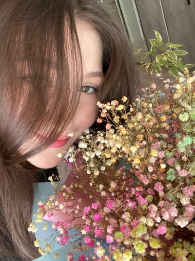 A young woman is holding a bouquet of multicolored dried baby's breath flowers. Her face is partially obscured by her brown hair, with her right eye and lips visible. The flowers are a mix of pink, white, yellow, blue, and green. The background is out of focus, but appears to be an indoor setting with hints of blue and pink packaging or material behind the flowers. The image is a close-up portrait taken from a slightly high angle. There are no identifiable landmarks or elements that would indicate a specific location within Nizhnyaya Maktama, Russia.