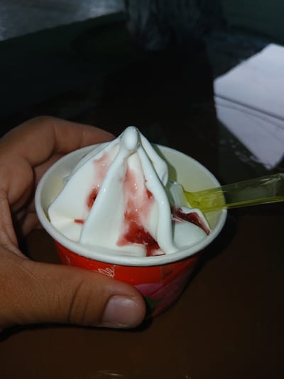 A hand holds a red paper cup filled with white soft-serve ice cream swirled with streaks of red strawberry syrup. A yellow plastic spoon rests on the rim of the cup, its handle extending to the right. The background is dark and out of focus, with a hint of a white rectangular object on the right side. The scene is likely indoors, potentially in a casual setting like a cafe or at home in Bazar-Korgon, Kyrgyzstan.