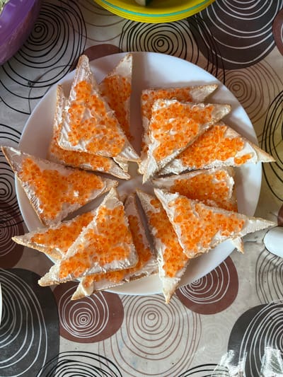 A white plate holds a pile of triangular white bread toast triangles topped with cream cheese and red caviar. The plate rests on a patterned tablecloth with concentric circles. The setting appears to be an indoor environment in Myrne, Ukraine, possibly a dining area or kitchen.