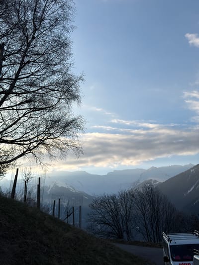 The image shows a mountain landscape near Saint-André, France. A truck is parked on the right side of a road. Silhouetted bare trees line the edge of the road. Behind the trees, there is a fence and a grassy slope. Snow-covered mountains are visible in the background with thin clouds. The sky is a light blue.
