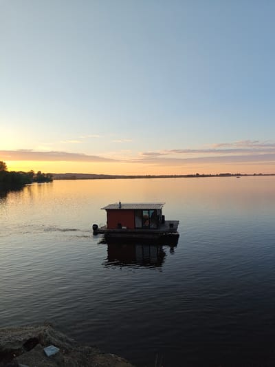 A wide-angle, vertical photograph captures a tranquil scene on a large body of water at either sunrise or sunset, specifically in the area of Nikolo-Beryozovka, Russia. The sky transitions from a pale blue at the zenith to a warm golden-orange and pink closer to the horizon, dotted with sparse, elongated clouds.
The central subject is a small, rectangular house-like structure, painted reddish-brown, situated on a dark floating pontoon or raft in the middle distance. This structure features a dark, shingled roof with a prominent silver chimney pipe. Large glass windows or doors are visible on the side of the building, reflecting the ambient light. A black Mercury brand outboard motor is attached to the stern of the pontoon, generating a discernible wake on the water's surface, indicating recent movement or current propulsion.
The water reflects the sky's changing colors, appearing golden-orange towards the left horizon and darker blue-grey in the foreground and to the right. Ripples cover the entire surface. To the left, a dense, dark green treeline marks the nearest bank, silhouetted against the glowing sky. Along the distant horizon, low hills and scattered trees are faintly visible, and at least one other small, dark boat can be discerned on the water in the far right distance.
The immediate foreground, at the bottom left, reveals a rough, earthy bank composed of dark, uneven rock or concrete debris, with a small, light-colored stone resting on it. No individuals are visible in the image.
