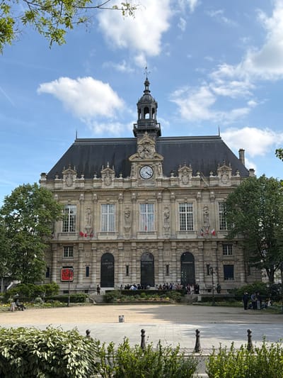 This media file captures the historic Hôtel de Ville (Town Hall) of Ivry-sur-Seine, France, under a clear, partly cloudy sky during the daytime. The imposing stone building features classical architecture with ornate carvings, multiple windows, and a prominent clock tower topped with a spire and weather vane. The dark, pitched roof is made of slate.

In front of the building is a public space with a paved area and a dirt path, flanked by green bushes, trees, and some flowering plants. Several people are visible in this area; some are sitting on benches, while others are walking or standing, creating a relaxed, everyday scene. Dark, decorative bollards line the edge of the path.

Notable details include small French flags hanging below some of the windows on the building. The clock on the tower reads approximately 2:10 or 2:11. Prominently displayed on the building's facade, likely on doors or plaques, are the French national motto words: "LIBERTE", "EGALITE", and "FRATERNITE". The text "HOTEL DE VILLE" is also visible on the building itself, confirming its identity. A red sign near the base of the building appears to bear the name "IVRY" as part of a larger text or logo, reinforcing the location in Ivry-sur-Seine. The overall atmosphere is calm and pleasant, indicative of good weather.