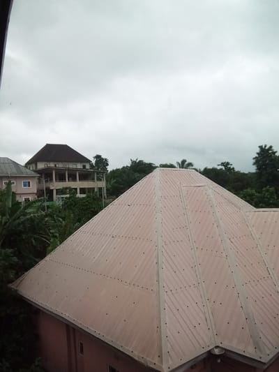 The image shows a view of several buildings and a predominantly overcast sky. In the foreground, a large, light brown metal roof with a corrugated texture dominates the lower half of the frame, extending diagonally from the bottom right to the center. This roof is secured with numerous dark-colored rivets. Adjacent to this is a portion of another building's roof, visible on the far left, which appears to have a gray, tiled surface and solar panels.
Behind the foreground roof, a multi-story building is visible. This structure has a light-colored facade and a dark, pyramidal roof. It features a wide, covered balcony or veranda supported by columns on the second floor. Further in the background, more buildings and dense green foliage, including banana trees and other tropical vegetation, are present. The sky is a uniform light gray, suggesting cloudy or overcast weather. The scene appears to be located in a residential area, possibly with a tropical climate, given the vegetation.
This location is identified as Nanka, Nigeria.