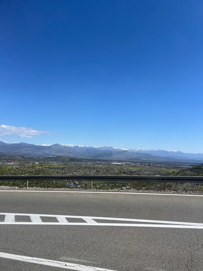 The image shows a view from a road in Gornji Kokoti, Montenegro, overlooking a valley with a town and mountains in the background. The sky is clear and blue. The road is paved with asphalt and has white lane markings. A metal guardrail runs along the edge of the road. The town in the valley appears to have houses with red roofs and patches of greenery. The mountains in the background are partially covered with snow.