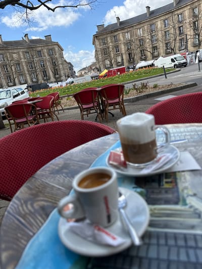 The image captures an outdoor cafe scene in Toul, France. Two cups of coffee, one a latte and the other an espresso, sit on a wooden table with a blue runner. The table is surrounded by red chairs, typical of outdoor cafes. In the background, buildings with multi-paned windows, flowerbeds, and parked white vans are visible. The sky is partly cloudy, suggesting a daytime setting. The perspective is slightly angled, focusing on the coffees while providing context of the city street.