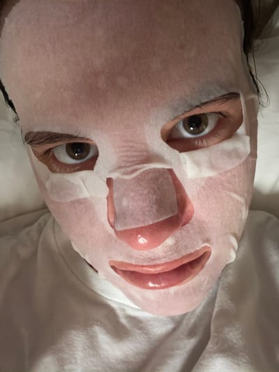 This media file shows a close-up, overhead shot of a person's face with a sheet mask applied. The person is lying down, likely in bed, as evidenced by the white, soft fabric of what appears to be bedding or a pillow underneath. The lighting suggests it could be daytime or an indoor setting with ambient light. The sheet mask covers most of the face, with cutouts for the eyes and mouth. The person's eyes are open and looking directly at the camera, displaying a warm, brown iris. Their lips are also visible through the mouth opening of the mask, appearing slightly glossy. The overall scene evokes a sense of relaxation and self-care. There is no visible text in the image. The location context provided is Vienne, France, but there are no specific visual cues in the image to confirm this location.