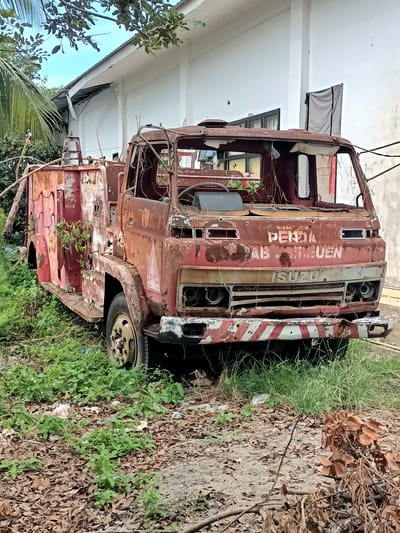 The image shows a rusted, abandoned fire truck in Blangbladeh, Indonesia. The truck is red and appears to be very old, with significant rust and deterioration. The windshield is broken, and vegetation is growing around and on the vehicle. The truck is parked on a dirt and leaf-covered surface, with green grass and trees surrounding it. A white building with a corrugated roof is visible in the background. The overall scene suggests neglect and the passage of time, with the fire truck being reclaimed by nature.