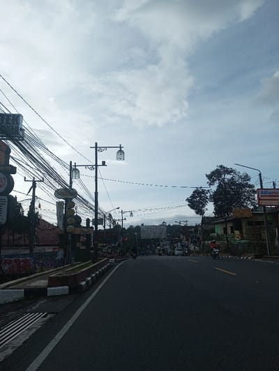 A street stretches into the distance under a cloudy sky. Buildings line the sides of the road, including a shop with an "Alfamart" sign. Power lines crisscross overhead, suspended from multiple utility poles adorned with decorative cage-like lanterns. Motorbikes travel on the road. A concrete barrier with alternating black and white blocks runs along the left side of the road, with graffiti visible on the adjacent wall.