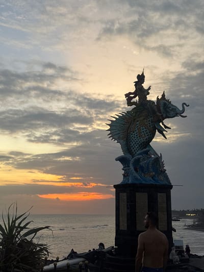 At sunset in Mengwi, Indonesia, a large, ornate statue of a mythical creature resembling a fish with an elephant riding on its back stands prominently. The statue is supported by a dark, multi-tiered base. A man, shirtless and wearing blue shorts, stands with his back to the camera, looking out at the ocean. The sky is filled with dramatic clouds, transitioning from soft yellow and orange hues near the horizon to darker gray tones above. The ocean is visible in the background, with gentle waves rolling towards a dark sand beach. Several other people are visible in the distance on the beach. Lush green vegetation is present in the foreground to the left of the frame.