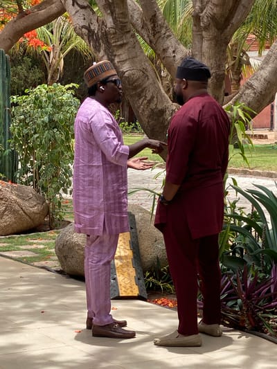 The image shows two men engaged in conversation outdoors, likely in a garden or courtyard setting in Kwarau, Nigeria, under bright daytime conditions.
The man on the left wears a lavender-colored traditional outfit with a subtle pattern, a multi-colored striped cap, and dark sunglasses. White wireless earbuds are visible in his ears. He stands facing the right, with his right arm raised and hand open, seemingly mid-gesture during a discussion. He is wearing brown loafers with a metallic buckle.
The man on the right, dressed in a maroon traditional outfit and a black skullcap, stands facing the left. He appears to be looking down towards the other man's gesturing hand or the ground. He wears light-colored, possibly beige, slip-on shoes.
The setting is lush with abundant greenery, featuring large trees with rough bark, tropical fronds, and various green and purple leafy plants. Several large rocks are scattered on the ground, and a yellow and black textured mat or cable protector is visible near the man in lavender. Both men are standing on a light-colored paved surface, with noticeable shadows indicating strong sunlight. No discernible text is visible in the image.