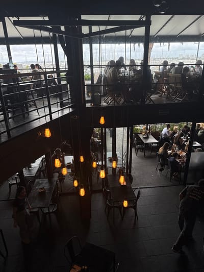 The image depicts a dynamic scene within a restaurant or bar, characterized by a striking contrast between its dimly lit industrial interior and a bright outdoor terrace overlooking a marina. The viewpoint is from an elevated position within the dark indoor area, looking out through large glass panels.
Inside, the decor features dark metal structures, tables, and chairs. Numerous Edison-style light bulbs hang at various heights, casting a warm, amber glow that illuminates the immediate surroundings and creates a cozy, modern ambiance. A person is seen walking across the dark tiled floor in the foreground.
Beyond the expansive windows, an outdoor dining terrace is bustling with patrons seated at wooden tables. This area offers clear views of a marina filled with numerous boats docked in the water, indicating a waterfront location in Punta del Este, Uruguay. The sky appears overcast or partly cloudy, suggesting daytime conditions, possibly late afternoon. Reflections of the interior elements, such as tables and lights, are visible on the glass, merging the indoor and outdoor spaces visually.
People on both sides of the glass appear relaxed, engaged in conversation and dining, contributing to a lively social atmosphere. A noticeable detail is a sign with the word "SALIDA" (Spanish for "EXIT") positioned above an doorway leading to the outside area. The overall setting combines urban architectural elements with a picturesque natural backdrop, highlighting the establishment's prime location.