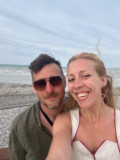 A couple takes a selfie on a pebble beach in Saint-Valery-en-Caux, France. The man, on the left, wears sunglasses and an olive-green shirt. The woman, on the right, smiles widely, her blonde hair blowing in the wind. She wears a cream-colored top with red trim. The sea and sky are visible in the background.