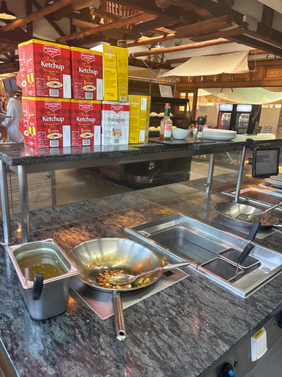 This is a vertical, medium-shot photograph taken inside a restaurant or food service establishment, likely in Gebersdorf, Austria. The scene depicts a food preparation or serving area with a focus on bulk condiments.
In the foreground, stacks of red boxes labeled "MAUTNER MARKHOF Ketchup" and yellow boxes labeled "FELIX MAYONNAISE" are prominently displayed on a counter. The ketchup boxes have images of tomatoes and are marked with expiration dates. To the right of the ketchup boxes, a stainless steel deep fryer is visible, containing two metal baskets. Further to the right, a large wok-style pan sits on a warming element, containing cooked food with vegetables and a serving utensil. A smaller metal container holding a yellowish liquid, possibly a sauce or broth, is next to the wok.
In the background, the setting appears to be a rustic restaurant with wooden beams and paneling. A steering wheel, possibly part of a decorative element, hangs on the wooden structure. A small bottle of TABASCO Original Pepper Sauce is visible on the counter near some white bowls and salt and pepper shakers. A digital tablet is mounted on a stand above the deep fryer, displaying a screen with what appears to be a menu or order interface. The overall impression is that of a busy food service environment, possibly during operating hours, given the prepared food and readily available condiments. The lighting suggests it is daytime, but no specific time can be determined. The image captures a utilitarian aspect of food service operations.
