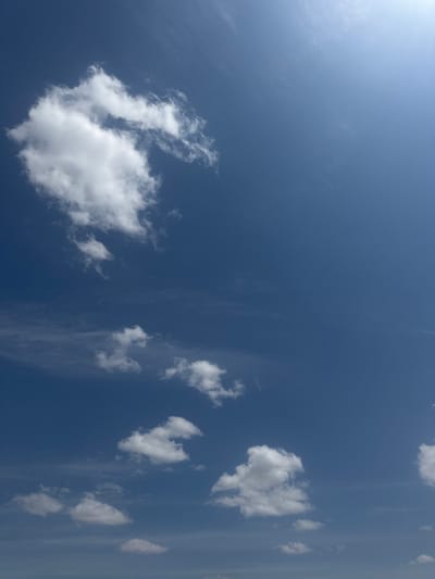 The image shows a clear blue sky with scattered white clouds. The sun is visible in the upper right corner, casting a bright light across the scene. There are no people, buildings, or other objects visible in the image, indicating an open sky view, possibly from an outdoor location. The environment appears to be natural and atmospheric, with no specific urban features from село Умбеталы Карибаева, Kazakhstan being depicted.