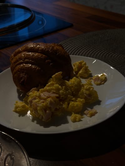 This media file shows a close-up, overhead view of a white plate on a dark wooden table, likely in a home or restaurant setting in Słupsk, Poland. The plate contains a breakfast meal consisting of scrambled eggs with small pieces of ham, and a croissant. The lighting suggests it may be early morning or evening, as harsh shadows are cast across the plate and surrounding table. The scene is dimly lit, with the main light source coming from the upper right, highlighting the food and the rim of the plate. The background shows a dark, textured placemat or tablecloth and the grain of the wooden table. To the left of the plate, a sliver of a reflective surface, possibly a glass tabletop or a futuristic-looking appliance, is visible with a blueish hue. No text is visible in the image. The focus is entirely on the food, presenting a simple yet appetizing breakfast.