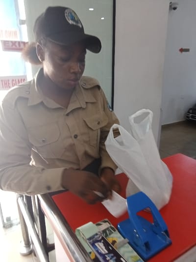 The image shows a young woman wearing a tan uniform, a black cap with a badge, and black pants. She is standing behind a counter, working with white plastic bags. In front of her is a blue hole punch on a red surface. The counter has a silver metal bar in front of it. There is a glass door in the background. The location, based on the provided coordinates, is near the intersection of Market Road and Emeka Anyaoku Street in Sapele, Nigeria. The scene is a retail establishment, possibly a store or a market stall.