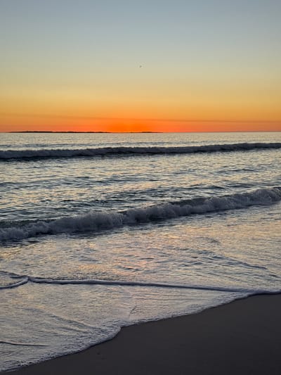 This high-angle shot captures a serene coastal scene at Blouberg, South Africa, likely during sunrise or sunset. The image, though oriented vertically, depicts a horizontal vista where the sky meets the ocean at a distinct horizon line.
The sky, occupying the upper portion of the frame, displays a warm gradient of colors, transitioning from vibrant orange and red tones directly above the horizon to softer yellows and pale blues higher up. A solitary, dark, bird-like shape is faintly visible against the lighter sky.
Below the horizon, the ocean stretches outwards, characterized by several parallel lines of waves. Closer to the viewer, the waves are breaking and receding over a dark, wet sandy beach. The intricate patterns of the foam and wet sand are highly detailed, showcasing the texture of the moving water and the shoreline. The wet sand on the right side of the image appears darker, contrasting with the foamy white edges of the waves.
The scene is tranquil and devoid of human presence or structures, focusing purely on the natural elements of the beach and ocean under a colorful sky. No text is visible in the media file.