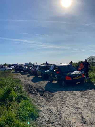 The image shows a line of racing cars parked on a dirt track, likely in Kanjiza Municipality, Serbia, judging by the Cyrillic text on the car. The cars are a mix of colors and models. Two people are leaning into the front car on the right, which is black with orange details. The area around the track has green vegetation. The sky is blue with streaks of clouds and a bright sun.