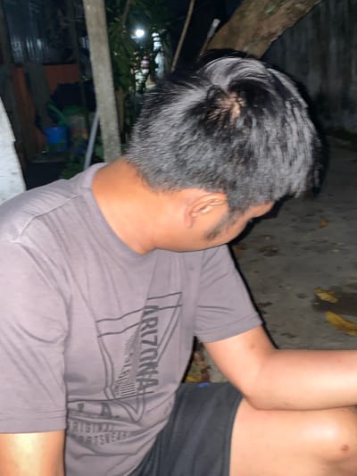 The image shows a person from behind, with their head turned down and to the side. They are wearing a grey t-shirt with a graphic design on it and black shorts. The person is outdoors, with a concrete ground visible, along with some fallen leaves and what appears to be a tree trunk in the background. There are also some objects and structures in the background that suggest a cluttered, possibly storage area, with a corrugated metal fence visible. The lighting suggests it is nighttime or dusk. The overall impression is a casual, informal setting. The specific location in Banjarmasin, Indonesia, cannot be determined from the visual information alone.