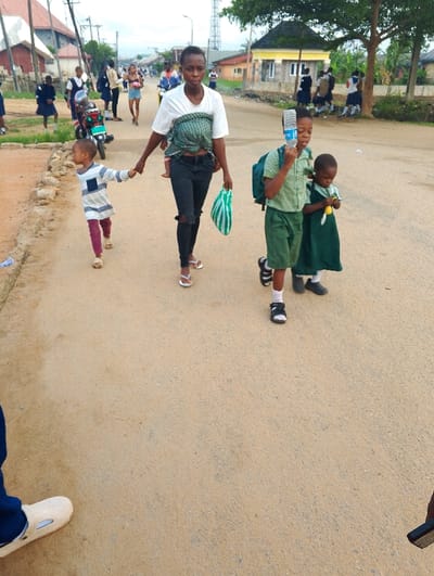 The image shows a group of people walking down a dirt road in Sapele, Nigeria. The road is lined with houses and trees on either side, and there is a group of people standing on the side of the road. The scene is located in Sapele, Nigeria, at approximately 5.93 latitude and 5.671 longitude. A woman in a white shirt and black jeans is leading the group, holding the hands of two young children. One child is wearing a blue and white striped shirt and the other is wearing a green shirt and a green dress. The woman is carrying a green and white striped bag in her left hand. There is a large blue water bottle in the hand of the young boy in the green shirt. Behind the woman, there is a group of children, all wearing green shirts and dresses, walking in the same direction. There are other people in the background, some are walking, others are standing, and some are riding bicycles. The scene appears to be a typical street in Sapele, Nigeria. The image is likely taken at a midday time of day, as the sky is bright and the shadows are relatively short.