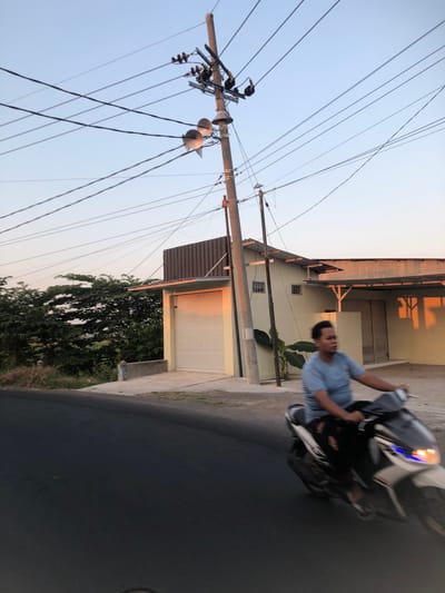 A person rides a motorcycle on a paved road in Glagah Arum, Indonesia, with utility poles and power lines overhead. The person, wearing a light blue t-shirt and dark pants, is in motion and their face is blurred. The motorcycle is white with some black and blue accents. To the right of the road is a light-colored building with a corrugated metal roof and a large garage door. In the background, there are dense green trees and vegetation. The sky is a pale blue, indicating either early morning or late afternoon. The scene is captured with a shallow depth of field, focusing on the motorcycle and rider.