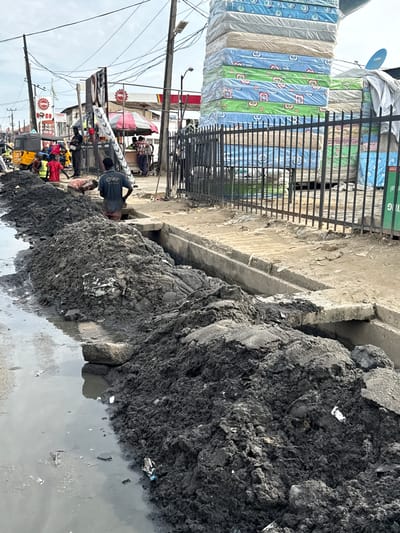 The image depicts a street scene in Lagos, Nigeria, characterized by significant urban infrastructure challenges. In the foreground, a large pile of dark, wet mud and debris has been excavated and deposited alongside a flooded drainage ditch. The muddy water reflects the overcast sky, and scattered pieces of debris are visible within the muck.
Several individuals are present, seemingly involved in or observing the ongoing work. One person in the middle ground, wearing a dark t-shirt with "CHAVY" visible, is standing near the drainage ditch. Another person, with their back to the camera, is bent over and appears to be working within the ditch. In the background, a yellow auto-rickshaw, a common mode of transport in Lagos, is parked, with people gathered around it.
The street is lined with various businesses and structures. Signs for "HIPCO" are visible, indicating a commercial presence. Further back, a stack of mattresses, wrapped in plastic, is piled high behind a black metal fence. The overall environment suggests a busy, working-class urban area. Overhead, a complex network of electrical wires and poles crisscrosses the sky, a common sight in many parts of Lagos. The scene conveys a sense of ongoing development or maintenance work, with the associated disarray and disruption to daily life.