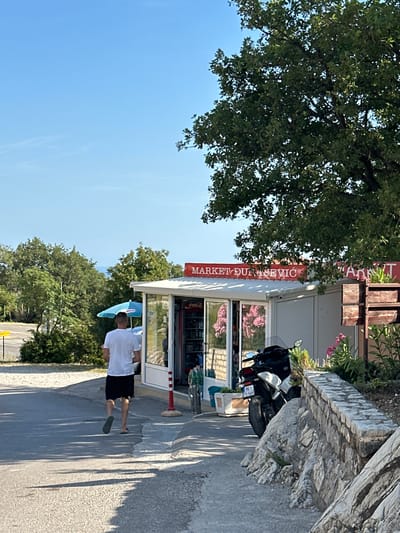 A man wearing a white t-shirt and black shorts walks away from the camera on a paved road in Blizikuće, Montenegro. To his right is a small market building with a red sign that reads "MARKET DULASEVIC". In front of the market, a light blue umbrella is open, and a motorcycle is parked. The market has glass windows displaying various items, including refrigerators stocked with drinks. Pink flowers are visible outside the market. The scene is set against a backdrop of lush green trees and a clear blue sky. The road is lined with a stone wall on the right side, and there are shadows cast on the road from the trees.