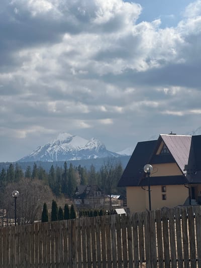 The scene depicts a panoramic view in Murzasichle, Poland, under a partially cloudy sky with a mix of grey and white cumulus clouds. In the foreground, a weathered wooden picket fence, composed of vertical planks, extends across the lower third of the image.
Behind the fence, to the left, a black, ornate streetlamp with a spherical glass globe is visible. Further back, a line of slender, dark green coniferous trees stands in front of a denser forest composed of both bare deciduous and evergreen trees. In the mid-ground on the right, a multi-story building with light tan stucco walls and a dark, corrugated metal A-frame roof is partially visible, featuring a small dormer window on its gable. Another identical streetlamp is partially obscured in front of this building. Several other buildings with similar architectural styles are interspersed within the forested landscape further back.
The background is dominated by a range of snow-capped mountains, with a prominent central peak heavily covered in white snow. A dense evergreen forest line forms a visual transition between the village structures and the mountain range. No individuals or active interactions are observed.
