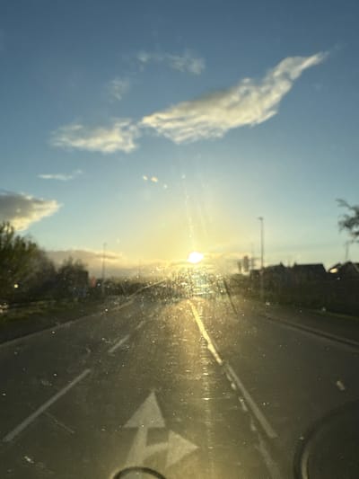 The image depicts a view from inside a vehicle, likely a car, looking out onto a road bathed in bright sunlight. The scene is oriented vertically. The sky is blue with scattered clouds, primarily visible on the left side of the image. The sun is positioned directly ahead, casting a strong glare and obscuring details in that area. The road is visible on the right, marked with white lines and arrows indicating traffic direction. The dashboard of the vehicle is partially visible at the top and bottom right. Vegetation lines the sides of the road, and buildings are seen faintly in the distance. The specific location in Oswestry, United Kingdom cannot be determined from the image.