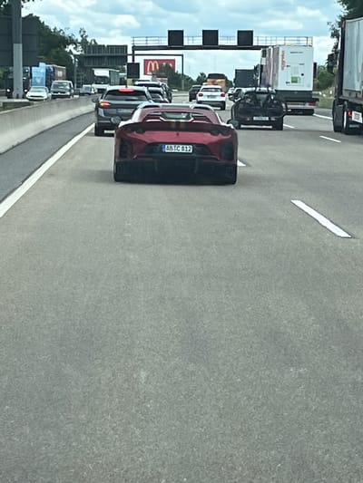 The image shows a rear view of a red sports car on a highway in Germany. The car has a license plate that reads "AB IC 812". Other vehicles, including trucks and SUVs, are visible on the highway. The scene appears to be in or near Klein-Krotzenburg, Germany, given the context. The sky is cloudy. The road surface is asphalt, and there are lane markings.