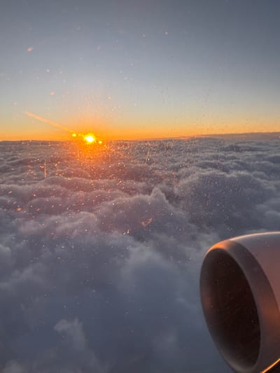 The image captures an aerial view from an airplane window during what appears to be either sunrise or sunset, presenting a dramatic sky and cloudscape. When viewed horizontally, the bright sun is a prominent feature on the left, glowing intensely with a golden-orange hue and creating a radiant streak across the frame. Its light brilliantly illuminates countless tiny specks and scratches on the airplane window, making them shimmer and sparkle across the foreground.
The sky above the sun transitions from a warm yellow to a soft, clear blue. Below and to the right of the sun, a vast expanse of textured clouds fills the mid-ground. These clouds are touched with warm orange and golden light on their upper surfaces where they catch the sun, while deeper grays, blues, and purples dominate their shadowed undersides and the clouds extending into the far right. A portion of the airplane's engine cowl is visible in the upper right corner, appearing dark but with its edges reflecting a warm, golden light.
The scene depicts an airplane in flight, high above a cloud layer, experiencing the "golden hour." The atmosphere is serene and expansive, with the primary activity being the journey itself. There is no visible text in the image. This captivating view provides a glimpse of the world from above, possibly during a flight near Vecsés, Hungary.