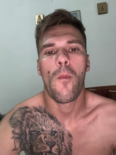 The media file is a close-up, low-angle, rotated portrait of a man. He has short, dark hair, brown eyes, and stubble. He is shirtless, revealing a large tattoo of a lion on his chest and shoulder. In the background, on a light-colored wall, there are two small framed pictures hanging. One appears to be a square wooden plaque with a depiction of a dog, and the other is a rectangular frame containing what looks like a drawing or photograph. The lighting suggests it is likely indoors and possibly nighttime, given the artificial light source. No specific weather conditions are discernible. The man's expression is neutral. There is no visible text in the image. The provided location context is Sesto Calende, Italy.