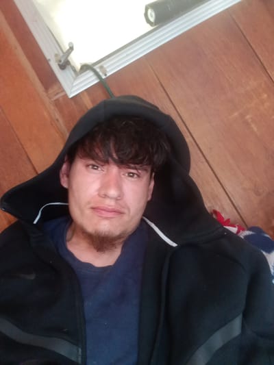 A young man is positioned centrally in a close-up, upward-angled shot. He has dark, wavy hair with bangs and a short goatee. He wears a dark blue t-shirt under a black zip-up hoodie, with the hood pulled up. A faint white stripe is visible along the edge of the hood, and a subtle Nike swoosh logo can be seen on the left sleeve of the hoodie. His expression is neutral to slightly contemplative as he looks directly towards the camera.
The background primarily consists of reddish-brown wooden paneling, suggesting an indoor setting like a room or cabin. In the upper left portion of the image, a segment of a white-framed structure, possibly a window or skylight, is visible. A metal latch mechanism and a dark green cord or wire are attached to this frame. A black, cylindrical object, resembling a flashlight, rests on the white frame in the upper right. In the bottom right corner, a glimpse of a patterned fabric, featuring red, white, and blue colors, is visible, possibly a blanket or throw. The overall impression is an informal portrait taken indoors.
