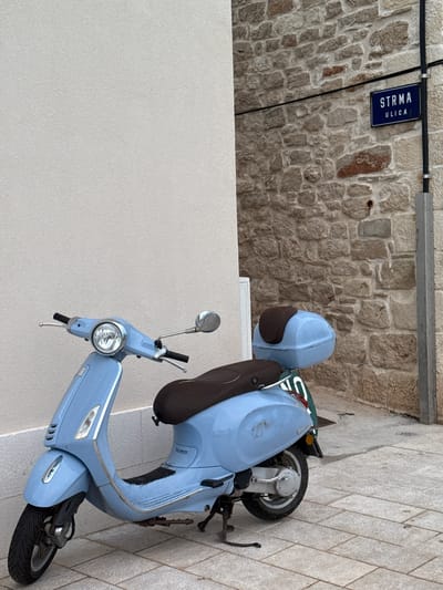 A light blue Vespa scooter is parked on a paved pathway next to a rough stone building in Primošten, Croatia. The scooter is facing left and has a brown seat and a matching top case. A street sign above the scooter reads "STRMA ULICA". The pathway is made of light gray rectangular stones. The building has walls made of rough, light brown stones with visible mortar. The sky is not visible in the frame.