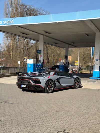 The image captures an Aral gas station in Adenau, Germany, under a clear blue sky. A grey Lamborghini Aventador SVJ Roadster, detailed with black accents and red pinstriping on its rear diffuser, side skirts, and wheel rims, is prominently parked on the light grey interlocking paver forecourt. The vehicle's German license plate displays "GL PV1". The gas station features a blue and white canopy with a corrugated underside, supported by white pillars. Underneath the canopy, multiple blue Aral fuel pumps are visible, some branded "Ultimate". To the far right, near the edge of the canopy, two motorcycles are parked; one is primarily white and black, the other is orange-red and black. Two individuals stand near the motorcycles; one wears a high-visibility yellow jacket over dark clothing, the other is partially obscured and dressed in dark attire. The background consists of a dense area of leafless deciduous trees and a distant building with a reddish roof, indicative of late winter or early spring. A metal guardrail is visible to the left, alongside a street light.