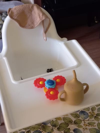 A high-angle medium shot captures a white plastic baby high chair with a bib draped over the top. On the tray of the high chair, there are three colorful plastic toys: a red and yellow flower-shaped toy, a blue and yellow toy, and a beige sippy cup. The background of the image is blurred, showing a dark wooden floor and a dark piece of furniture. The scene is indoors and appears to be in a home environment, possibly a kitchen or dining area. There is no indication of the location being in Baden, Austria.