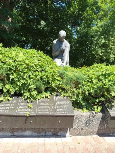 The image depicts a monument with a statue of a person kneeling, set amidst dense green foliage. The base of the monument is made of dark granite and is inscribed with names in Cyrillic script, suggesting it is a memorial. The environment appears to be outdoors, with trees and sunlight indicating a daytime scene. The overall impression is one of solemn remembrance. The monument is located in the city of Ivankiv, Ukraine.